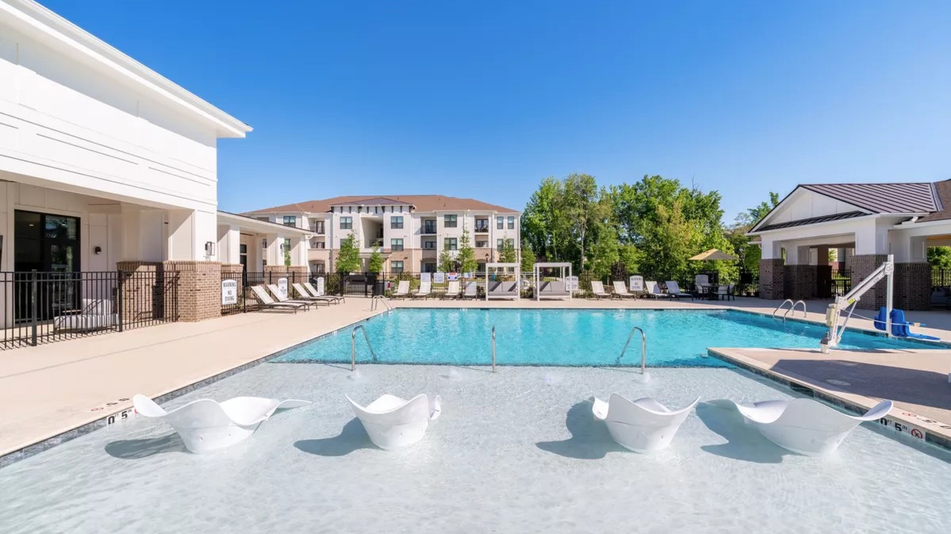 Outdoor swimming pool with white lounge chairs in shallow water, surrounded by apartment buildings and trees.