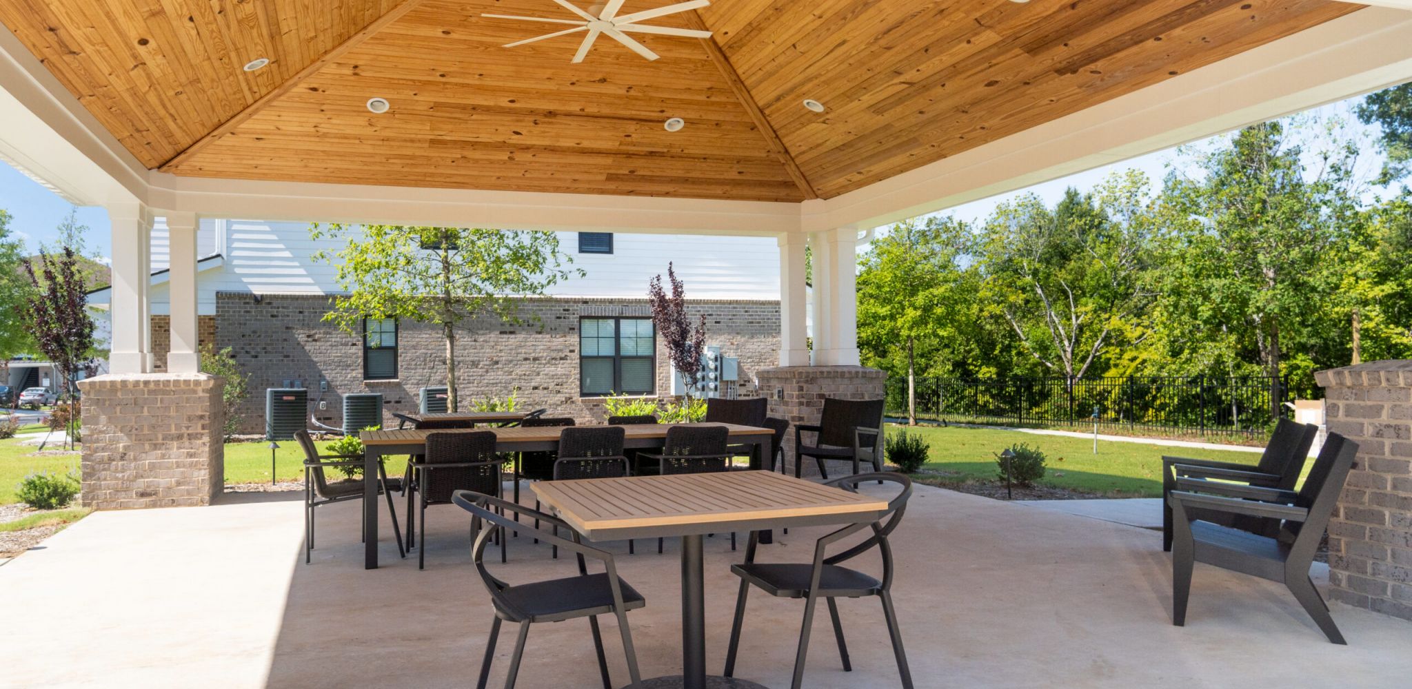 Covered patio with wooden ceiling, outdoor tables and chairs, and a view of a grassy yard with trees.